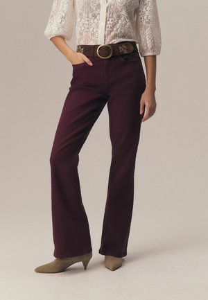 Nő bordó magas derekú, bővülő szárú nadrágban, barna övvel nagy csattal, törtfehér csipkés blúzban és bézs magassarkú cipőben, egyik kezét a zsebében tartva áll.