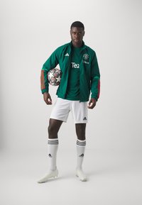 Chaqueta verde de adidas con rayas rojas, camiseta verde con el logo "Tezos", pantalones cortos blancos, calcetines blancos con rayas negras, sosteniendo un balón de fútbol en blanco y negro.