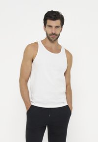 Man wearing a white sleeveless tank top and black pants standing against a plain white background with hands in pockets.