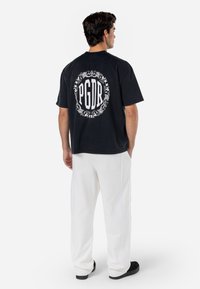 Black cotton t-shirt with large white circular graphic featuring "PGDR" and decorative details on the back. Paired with white pants.