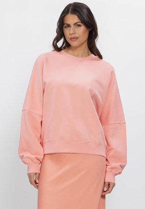 Frau mit langen dunklen Haaren, die einen lockeren rosa Pullover und einen passenden rosa Rock trägt, steht vor einem schlichten weißen Hintergrund.