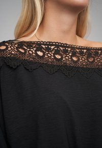 Svart topp med bar skuldre, prydet med intrikat svart blonderkant rundt halsen med blomsterkraft og bølgete kanter. Glatt tekstur.