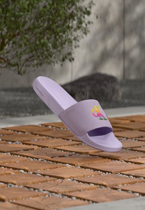 Lavendelfarvet Calvin Klein slide-sandal med flerfarvet "CK"-logo på rem, vist svævende over teksturerede udendørs fliser.
