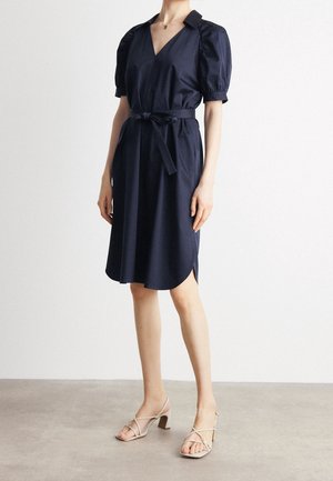 Kobieta w granatowej sukience do kolan z bufkami na rękawach, dekoltem w serek i paskiem zawiązanym w talerzu, w połączeniu z beżowymi sandałami na słupku z paskami.