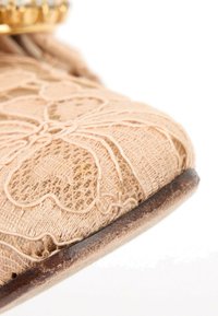 Gros plan du bout d’une chaussure beige recouverte de dentelle avec des motifs floraux complexes et une semelle marron usée sur fond blanc.