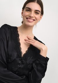 Blusa nera in satin con scollatura a V e bordo in pizzo, caratterizzata da maniche lunghe e dettagli con bottoni. Texture liscia con una silhouette aderente.