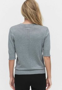 Kvinna med långt blont hår iförd en ljusgrå kortärmad tröja och svarta byxor, sedd bakifrån mot en enfärgad bakgrund.