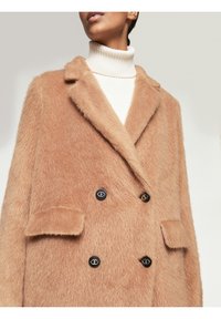Cappotto doppiopetto in tinta beige chiaro in pelliccia sintetica, dotato di un colletto a scollo, due tasche a toppa e bottoni neri con accenti dorati.