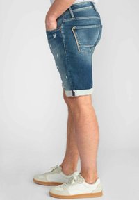 Leren denim shorts in vervaagd blauw met een versleten ontwerp, opgerolde zoom en vijf-pocket styling. Gepaard met witte sneakers met beige accenten.