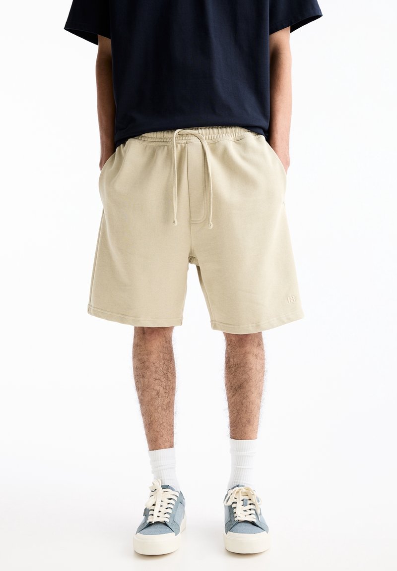 Personne portant un short beige avec cordon de serrage, des chaussettes blanches, des baskets bleues et un t-shirt noir, se tenant devant un fond blanc.