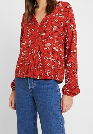 Femme portant un chemisier rouge à fleurs à manches longues avec des détails en dentelle, associé à un jean bleu, montrée du cou aux hanches.