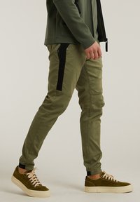 Pantalon extensible vert olive avec panneaux latéraux noirs, coupe fuselée et design minimaliste ; accompagné de baskets en daim marron avec lacets crème.