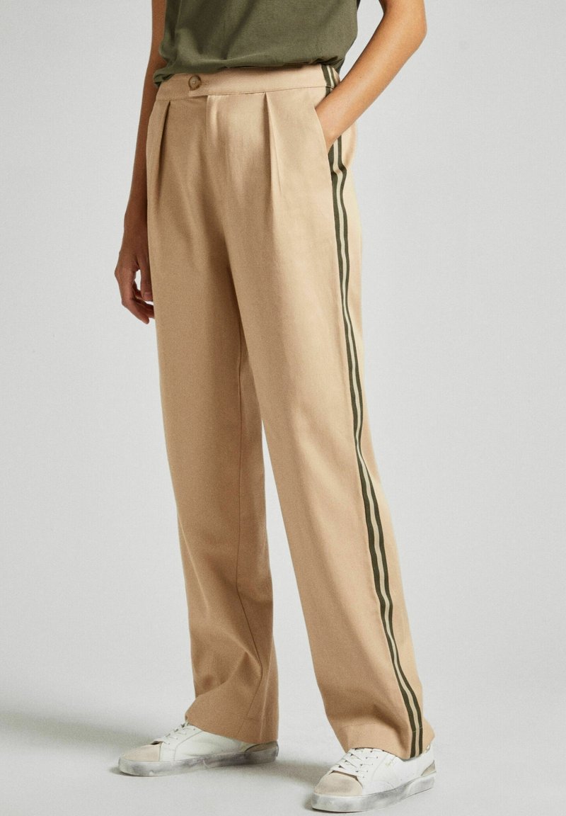 Pantalon large beige avec des bandes latérales foncées, associé à des baskets blanches et un haut vert, porté par une personne debout.