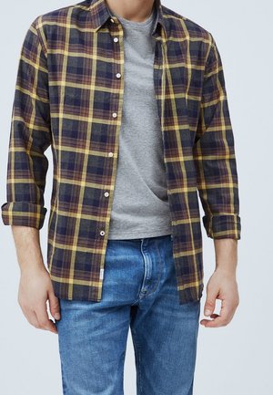 Camisa de franela en cuadros azul marino, amarillo y burdeos, con cuello y cierre de botones, superpuesta sobre una camiseta gris.