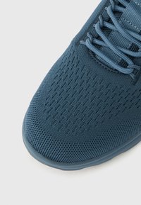 Κοντινή λήψη της μύτης ενός μπλε πλεκτού αθλητικού παπουτσιού με μπλε κορδόνια που ταιριάζουν και υφή στη σόλα από καουτσούκ, σε ανοιχτό φόντο.