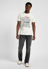 Camiseta de color crema claro con un gráfico de denim vintage en rojo y negro, combinada con unos jeans gris oscuro y zapatillas blancas con detalles en negro.