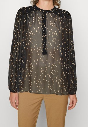 Musta läpikuultava paita, jossa on beige pilkullinen kuvio, pitkät puhvikäsivarret ja napitettu etulista, yhdistetty vaalean ruskeiden housujen kanssa.