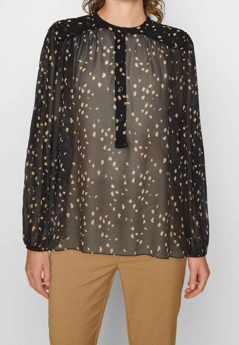 Blouse noire transparente à motif beige tacheté, manches longues bouffantes, patte de boutonnage à l'avant, portée avec un pantalon marron clair.