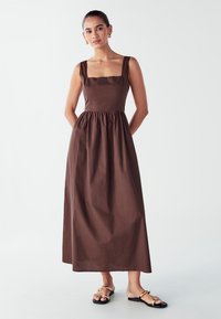 Rjava brezrokavna obleka s kvadratnim izrezom, zategnjenim pasom in polno krilo. Gladka tekstura, lahek material. Sandali z zlatimi detajli.