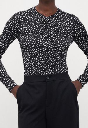 Osoba ubrana w dopasowaną czarną bluzkę z długim rękawem, ozdobioną białymi abstrakcyjnymi plamami, oraz czarne spodnie z wysokim stanem, trzymająca ręce w kieszeniach.