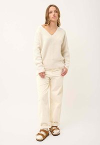 Cremfarbene Strickjacke mit V-Ausschnitt, kombiniert mit gerippten, cremfarbenen Hosen und braunen Sandalen. Weiche Textur, lockere Passform.