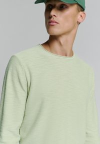 Lichtgroen shirt met lange mouwen, gemaakt van een textuurstof en een ronde hals, gecombineerd met een groene pet. Close-up van het bovenlijf.