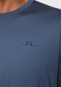 Blå atletisk skjorta gjord av glatt, fukttransporterande material. Har en subtil logotyp i en mörkare nyans på framsidan. Kortärmad design.