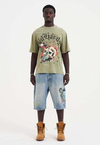 Oliivinvihreä graafinen t-paita, jossa on päähinekuvio, farkku-shortsit, joissa on värikkäitä kirjontatöitä, ruskeat nauhakiinnitteiset saappaat ja hopeakorut.