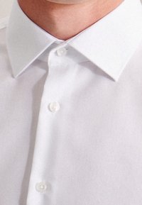 Camicia bianca in cotone con colletto classico a punta e una patta con bottoni perlati. Texture liscia con una finitura netta.