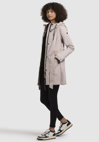 Veste imperméable rose clair, avec capuche, boutons-pression, cordon de serrage ajustable et poches plaquées, portée sur des leggings noirs et des baskets.