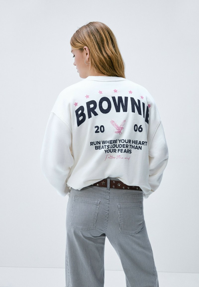 Sudadera blanca con estampado gráfico que dice "BROWNIE" y presenta un diseño de estrellas y un águila. Combinada con pantalones de rayas y un cinturón.