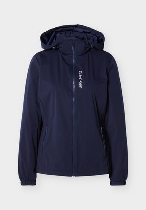 Ναυτικό μπλε μπουφάν με κουκούλα και φερμουάρ μπροστά, πλαϊνές τσέπες με φερμουάρ, ελαστικά μανίκια και κάθετο λευκό λογότυπο "Calvin Klein" στο αριστερό μέρος του στήθους.