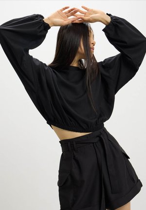 Felpa nera tagliata con orlo elasticizzato e maniche voluminose, abbinata a pantaloni corti neri sartoriali con cintura larga e tasche frontali.