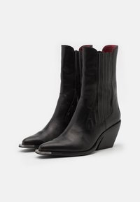 Stivaletti in pelle nera alla caviglia con punta affilata, bordino ondulato e pannelli laterali scanalati. Presentano un tacco a zeppa basso e accenti metallici.