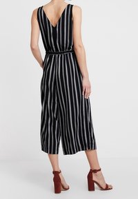 Combinaison à rayures noires et blanches avec un dos en V, sans manches et taille cintrée. Associée à des sandales à talons carrés marron.