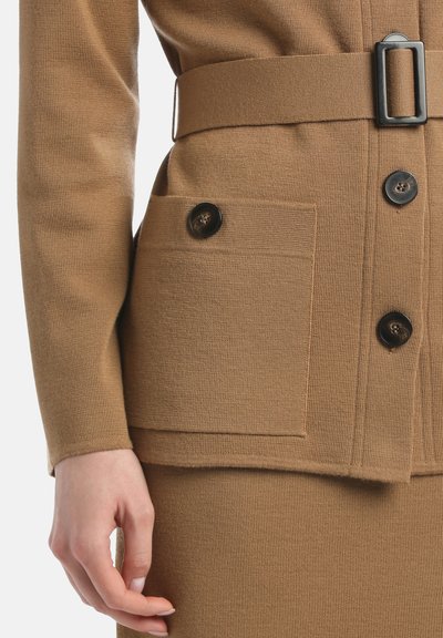 Gros plan sur une personne portant une robe en tricot marron avec une poche boutonnée et une ceinture assortie avec une boucle rectangulaire noire.