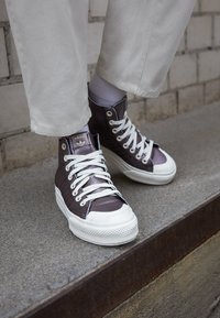 Höga sneakers i metallisk lila med vita gummisulor och snören. Texturerade tygpaneler och märkesdetalj på tungan synliga.