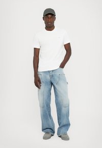 Vit t-shirt med rund halsringning, ljusblå baggy denimjeans och grå keps. Jeans med fickor och blekt utseende. Grå sneakers fulländar outfiten.