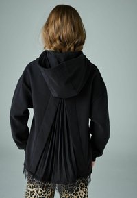 Čierna mikina s plisovaním na chrbte, zdobená čipkovým lemom na spodku. Modelka má na sebe legíny s leopardím vzorom.