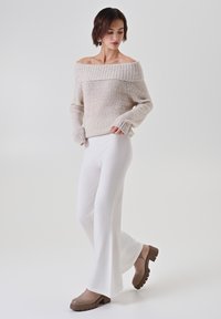 Maglione beige a spalla scoperta con scollatura a costine, abbinato a pantaloni bianchi svasati e sandali con tacco grosso marroni su uno sfondo neutro.