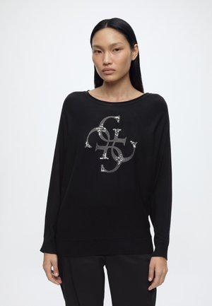 Jeune femme aux longs cheveux noirs portant un pull noir avec un logo abstrait orné d'argent sur le devant sur fond blanc.