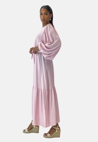 Vestido largo rosa con mangas voluminosas, cintura fruncida y dobladillo escalonado. Combinado con sandalias de cuña beige. Tela suave y ligera.