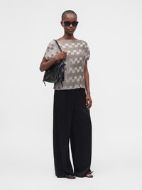 Femme aux cheveux courts portant des lunettes de soleil noires, un haut à manches courtes à motifs, un pantalon large noir, des sandales noires, et tenant un sac à bandoulière noir.