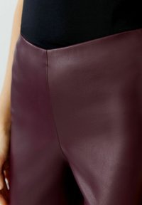 Burgunderfarbene Kunstlederhosen mit glatter Textur und geschwungenen Nähten, die zusammen mit einem schwarzen Oberteil präsentiert werden, wodurch der Bund betont wird.