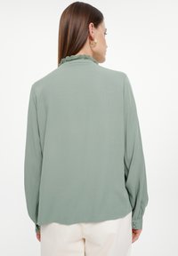 Blusa de manga larga, color verde claro, con cuello con volantes, puños abotonados y textura suave. Corte sencillo y relajado con dobladillo redondeado.