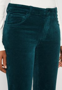 Turkosa kordflöjtsbyxor med vertikala ribbor, som har framfickor och en midja med bälteshällor. Mjuk textur och figurnära design.