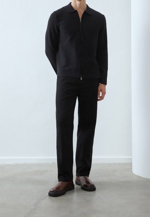 Pull noir zippé avec col, porté sur un pantalon noir. Chaussures marron à semelle épaisse. Texture lisse et design minimaliste.