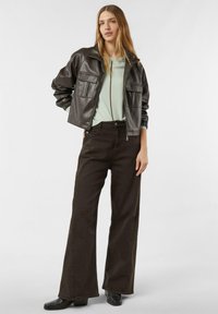 Kobieta ubrana w ciemnobrązową skórzaną kurtkę, jasnobeżowy sweter, szerokie ciemnobrązowe spodnie i czarne botki, stojąca na jednolitym tle.