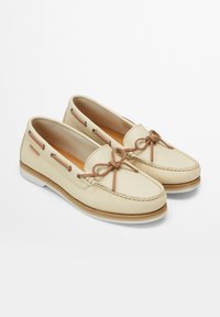 Beige mokasiner i läder med en strukturerad yta, utrustade med en båtdetalj, snöraccent och en vit gummisula med en träram.