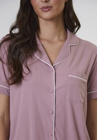 Camisa de pijama de manga corta y con botones en tejido de canalé de color púrpura claro. Presenta un ribete blanco en el cuello y en el bolsillo del pecho.